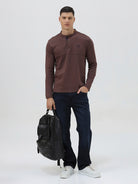A man wearing a brown long-sleeve henley shirt with a small embroidered logo on the left chest, paired with dark blue jeans, standing against a light gray background.