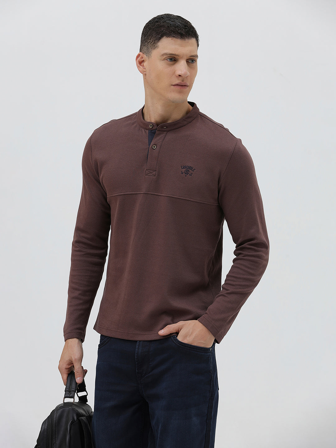 A man wearing a brown long-sleeve henley shirt with a small embroidered logo on the left chest, paired with dark blue jeans, standing against a light gray background.