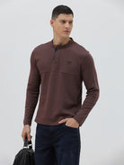 A man wearing a brown long-sleeve henley shirt with a small embroidered logo on the left chest, paired with dark blue jeans, standing against a light gray background.