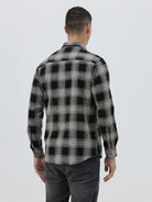 A man wearing a long-sleeve button-up plaid shirt in black, gray, and white, paired with dark gray jeans, standing against a light gray studio background.