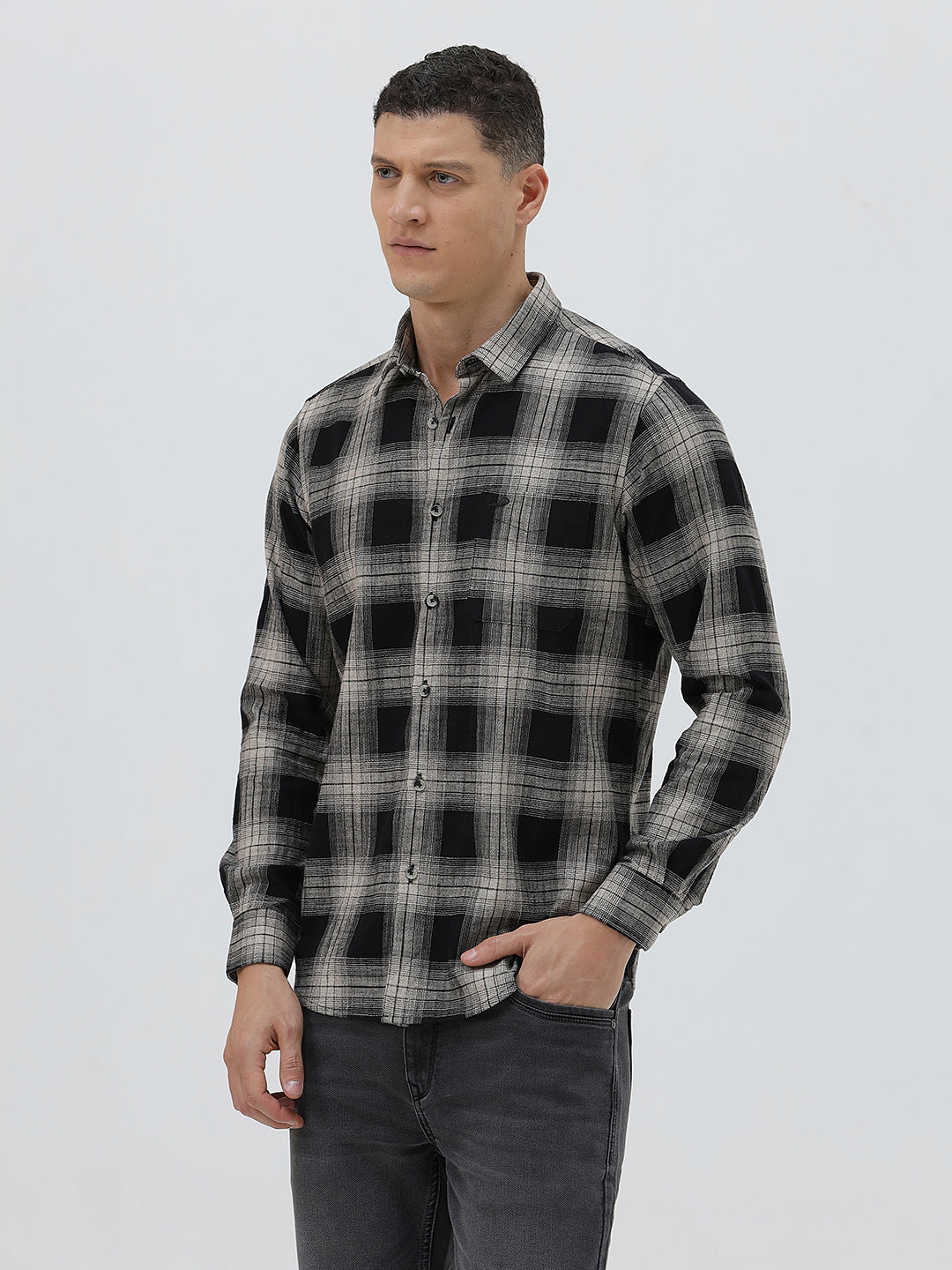 A man wearing a long-sleeve button-up plaid shirt in black, gray, and white, paired with dark gray jeans, standing against a light gray studio background.