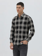 A man wearing a long-sleeve button-up plaid shirt in black, gray, and white, paired with dark gray jeans, standing against a light gray studio background.