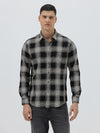 A man wearing a long-sleeve button-up plaid shirt in black, gray, and white, paired with dark gray jeans, standing against a light gray studio background.