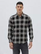 A man wearing a long-sleeve button-up plaid shirt in black, gray, and white, paired with dark gray jeans, standing against a light gray studio background.