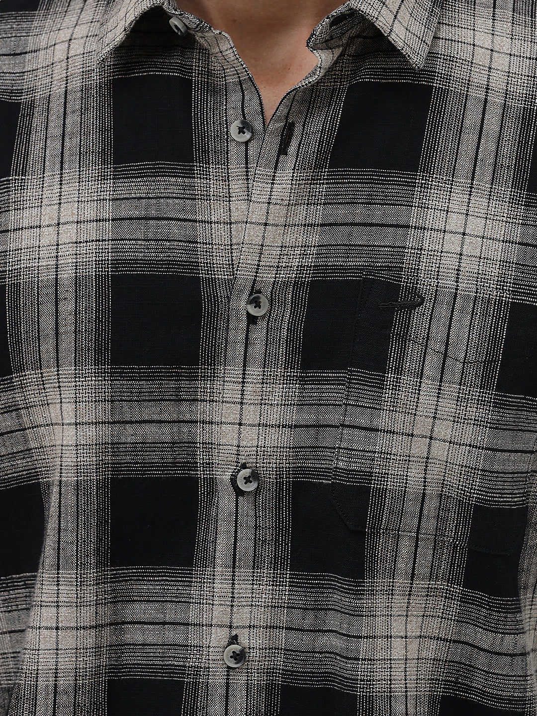 A man wearing a long-sleeve button-up plaid shirt in black, gray, and white, paired with dark gray jeans, standing against a light gray studio background.