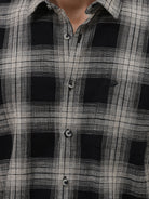 A man wearing a long-sleeve button-up plaid shirt in black, gray, and white, paired with dark gray jeans, standing against a light gray studio background.