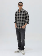 A man wearing a long-sleeve button-up plaid shirt in black, gray, and white, paired with dark gray jeans, standing against a light gray studio background.