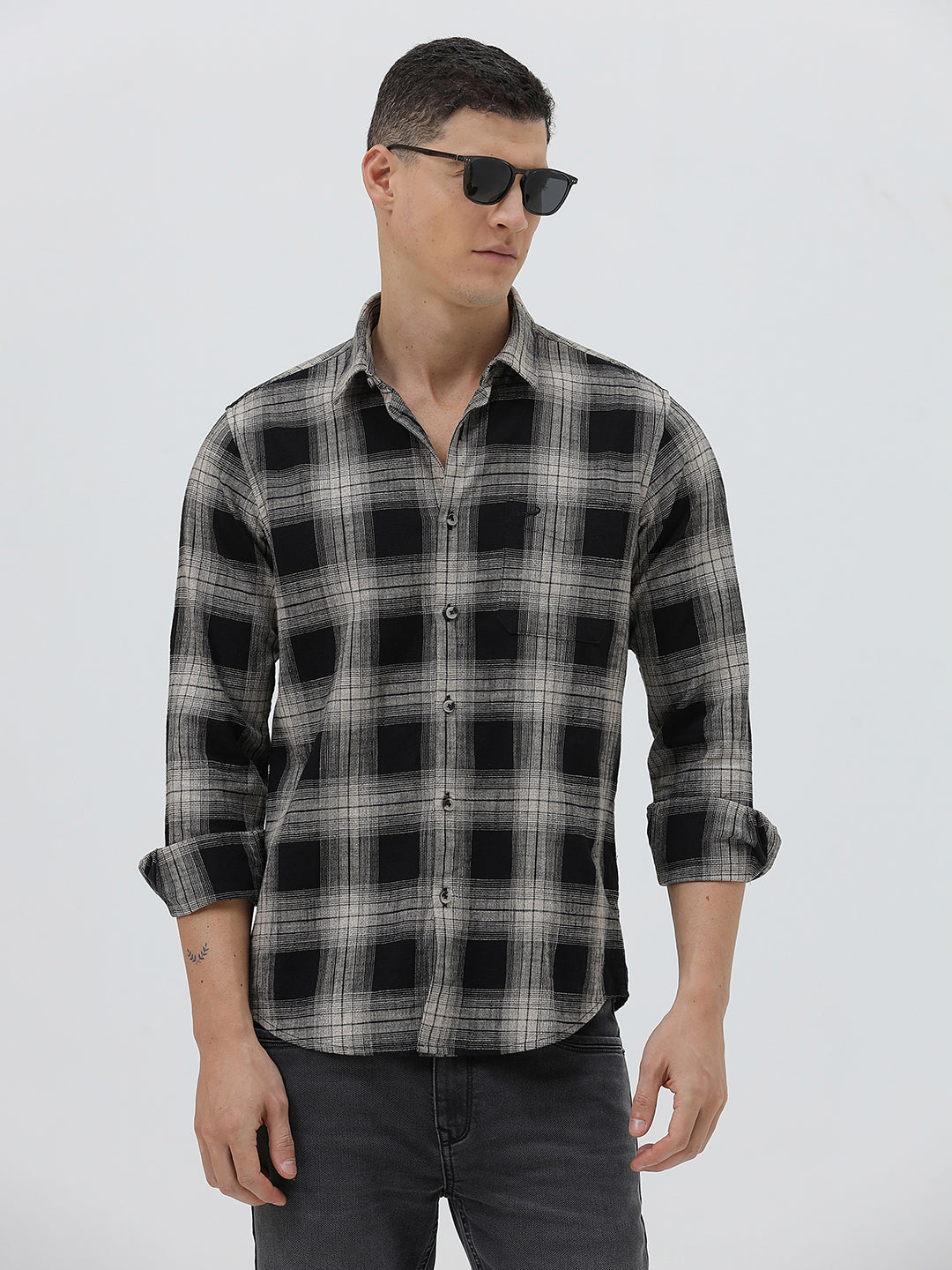 A man wearing a long-sleeve button-up plaid shirt in black, gray, and white, paired with dark gray jeans, standing against a light gray studio background.