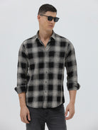 A man wearing a long-sleeve button-up plaid shirt in black, gray, and white, paired with dark gray jeans, standing against a light gray studio background.