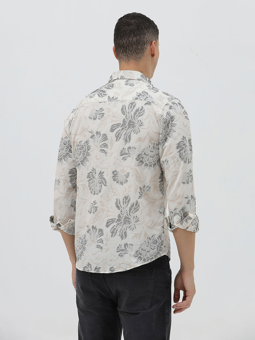 A man wearing a long-sleeve button-up shirt with a gray floral pattern on a cream-colored base, paired with dark gray trousers, against a plain light gray background.