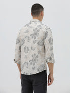 A man wearing a long-sleeve button-up shirt with a gray floral pattern on a cream-colored base, paired with dark gray trousers, against a plain light gray background.