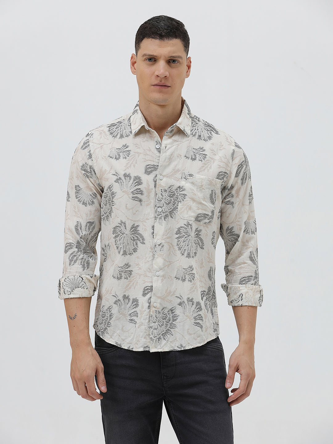 A man wearing a long-sleeve button-up shirt with a gray floral pattern on a cream-colored base, paired with dark gray trousers, against a plain light gray background.