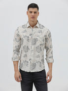 A man wearing a long-sleeve button-up shirt with a gray floral pattern on a cream-colored base, paired with dark gray trousers, against a plain light gray background.