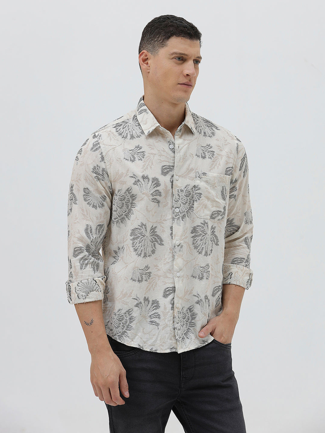 A man wearing a long-sleeve button-up shirt with a gray floral pattern on a cream-colored base, paired with dark gray trousers, against a plain light gray background.