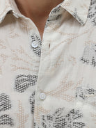 A man wearing a long-sleeve button-up shirt with a gray floral pattern on a cream-colored base, paired with dark gray trousers, against a plain light gray background.