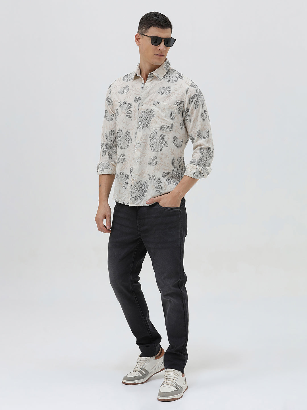 A man wearing a long-sleeve button-up shirt with a gray floral pattern on a cream-colored base, paired with dark gray trousers, against a plain light gray background.