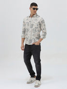A man wearing a long-sleeve button-up shirt with a gray floral pattern on a cream-colored base, paired with dark gray trousers, against a plain light gray background.