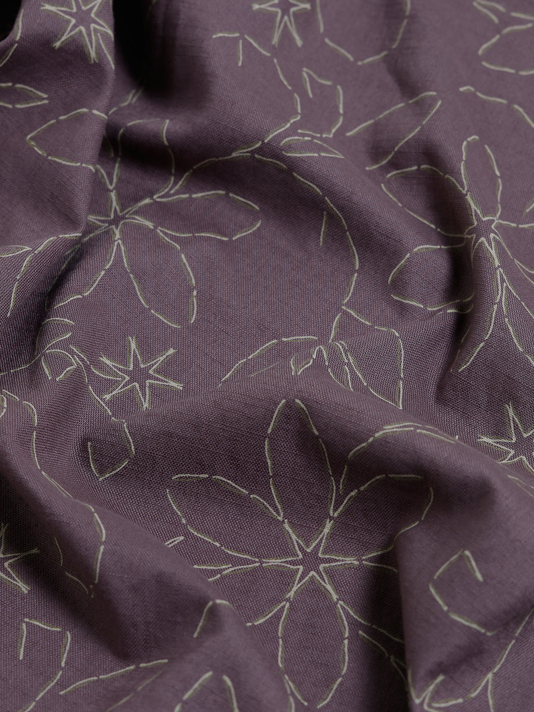 A man wearing a short-sleeve button-up shirt in a muted brownish-purple color with a light beige floral pattern, paired with khaki pants, against a plain light gray background.