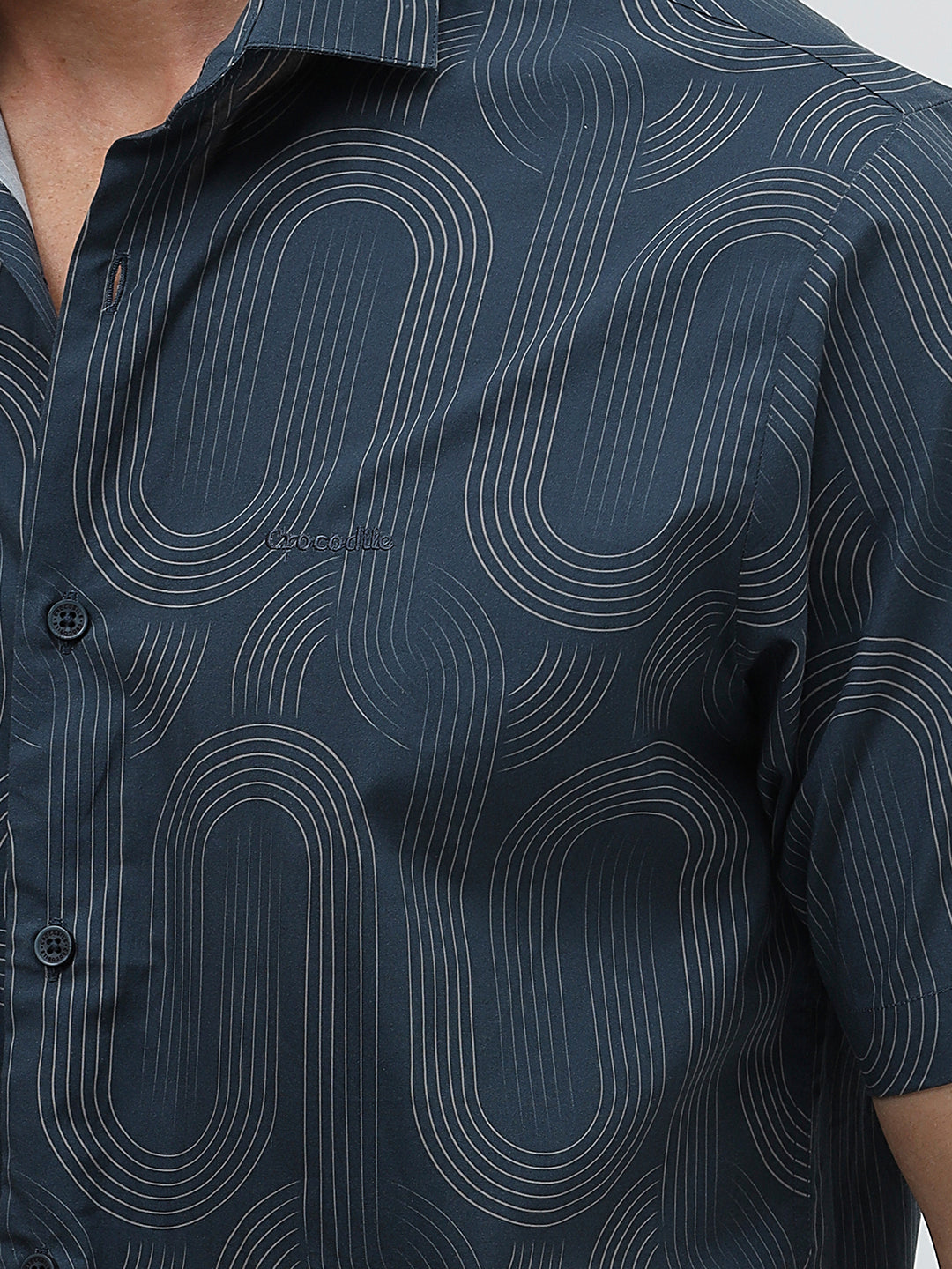 A man wearing a short-sleeve button-up shirt with a dark blue and gray abstract wavy pattern, paired with light beige trousers, standing against a plain light gray background.