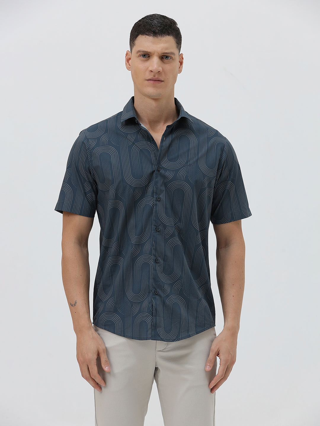 A man wearing a short-sleeve button-up shirt with a dark blue and gray abstract wavy pattern, paired with light beige trousers, standing against a plain light gray background.