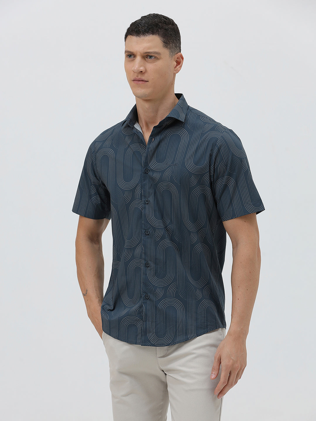 A man wearing a short-sleeve button-up shirt with a dark blue and gray abstract wavy pattern, paired with light beige trousers, standing against a plain light gray background.