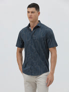 A man wearing a short-sleeve button-up shirt with a dark blue and gray abstract wavy pattern, paired with light beige trousers, standing against a plain light gray background.