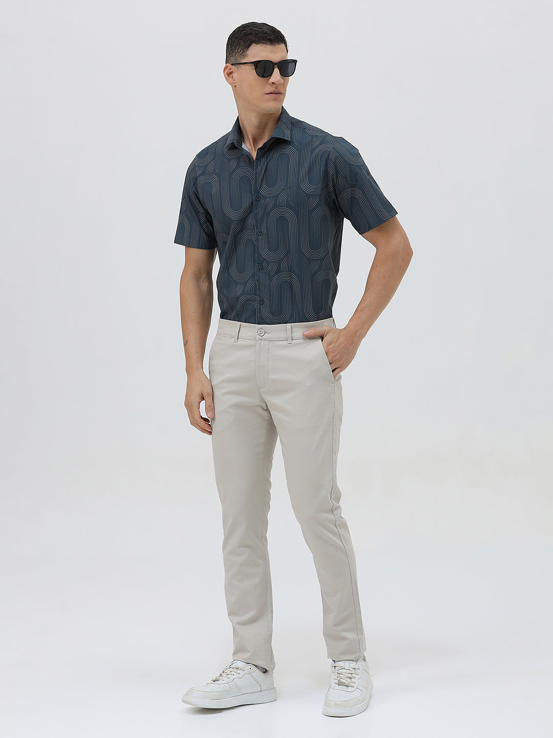 A man wearing a short-sleeve button-up shirt with a dark blue and gray abstract wavy pattern, paired with light beige trousers, standing against a plain light gray background.