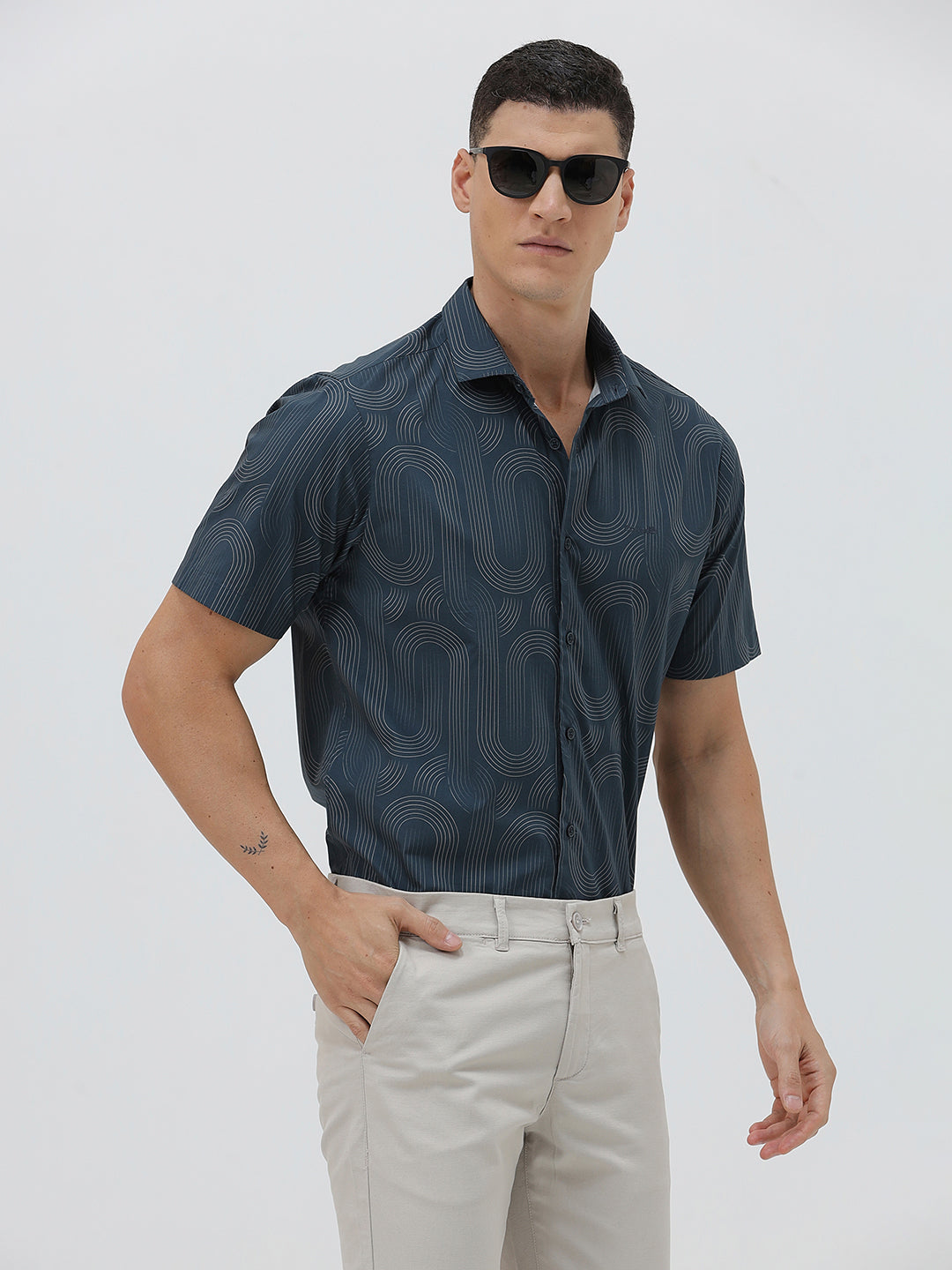 A man wearing a short-sleeve button-up shirt with a dark blue and gray abstract wavy pattern, paired with light beige trousers, standing against a plain light gray background.