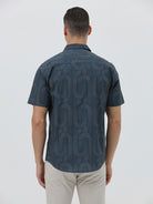 A man wearing a short-sleeve button-up shirt with a dark blue and gray abstract wavy pattern, paired with light beige trousers, standing against a plain light gray background.
