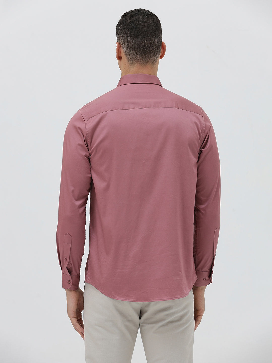 A man wearing a dusty rose long-sleeve button-down shirt and light beige trousers, standing against a light gray studio background.