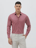 A man wearing a dusty rose long-sleeve button-down shirt and light beige trousers, standing against a light gray studio background.