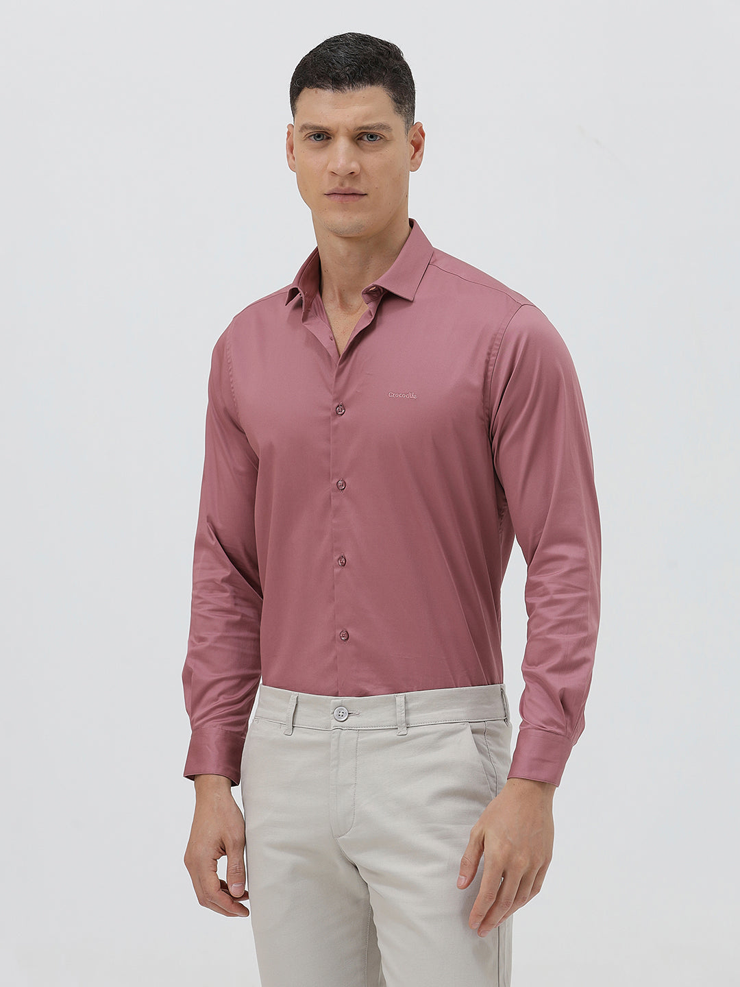 A man wearing a dusty rose long-sleeve button-down shirt and light beige trousers, standing against a light gray studio background.