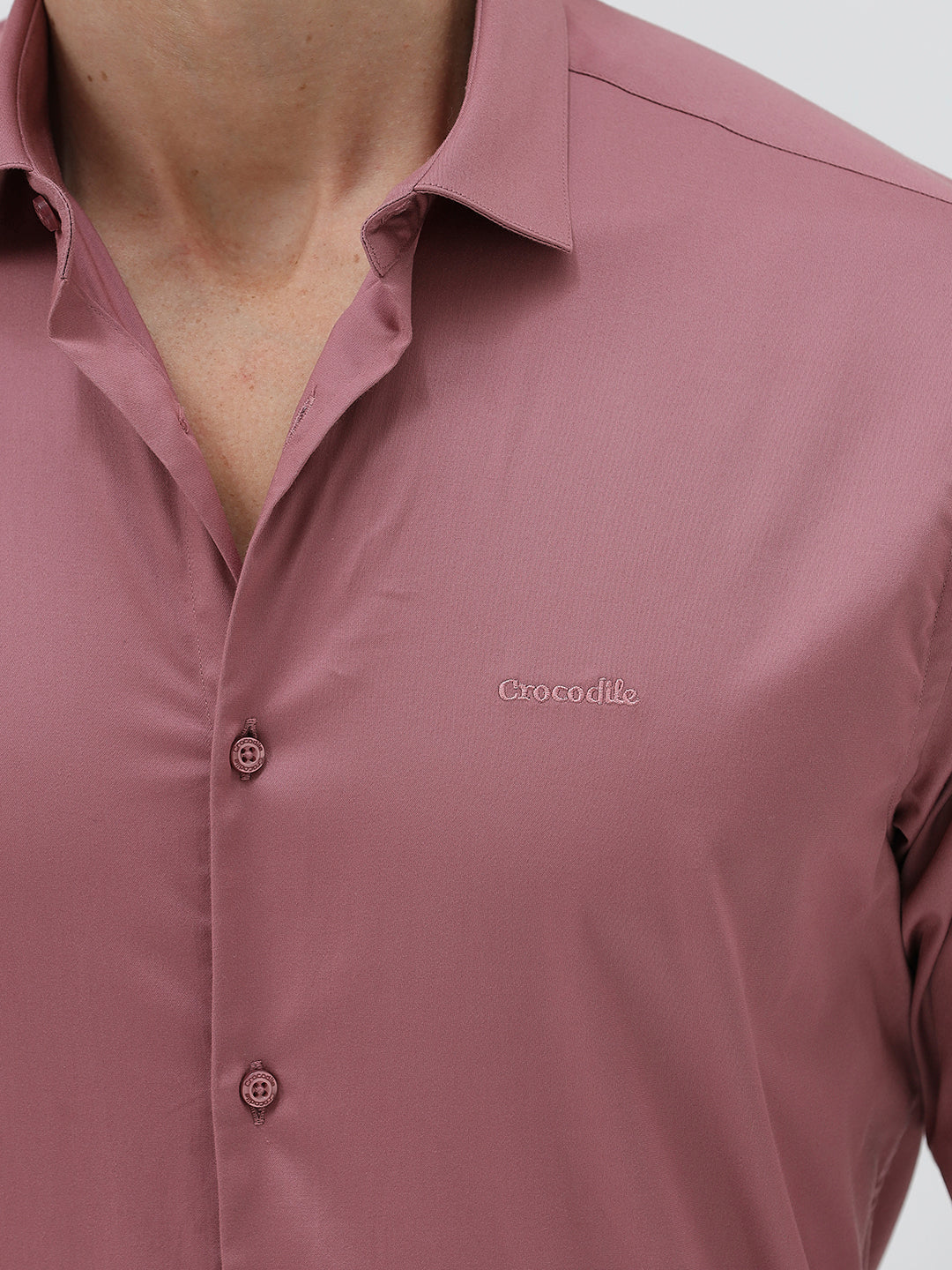A man wearing a dusty rose long-sleeve button-down shirt and light beige trousers, standing against a light gray studio background.