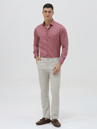 A man wearing a dusty rose long-sleeve button-down shirt and light beige trousers, standing against a light gray studio background.
