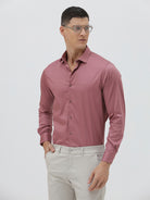 A man wearing a dusty rose long-sleeve button-down shirt and light beige trousers, standing against a light gray studio background.